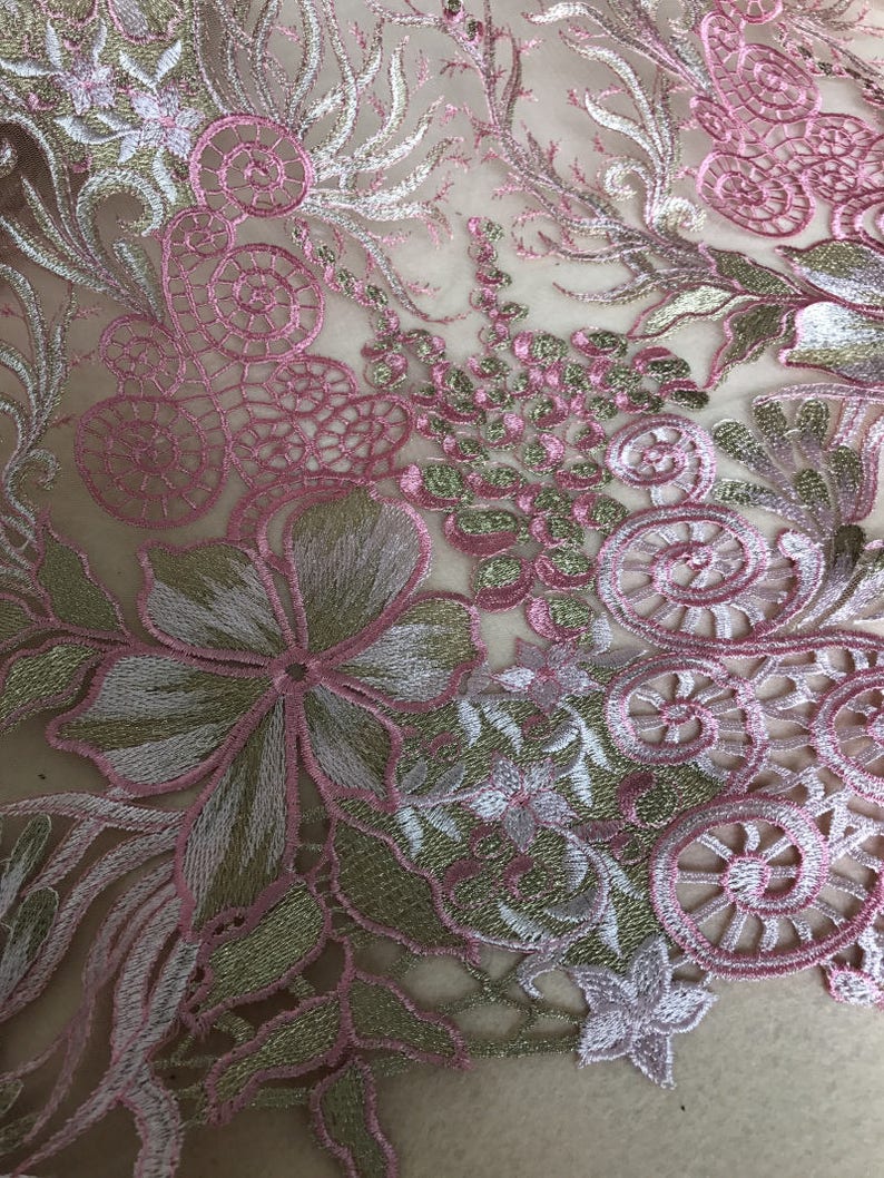 May include: Close-up of embroidered fabric featuring a floral design. The fabric has a sheer, light beige background with intricate embroidery in pink, gold, and white. The design includes flowers, swirls, and other decorative elements.