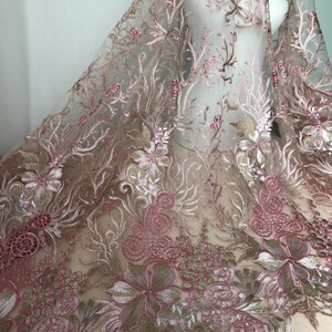 May include: Delicate pink floral embroidered lace fabric. The lace features intricate designs of flowers, leaves, and swirling patterns in shades of pink, white, and gold. The sheer fabric is draped, showcasing the detailed embroidery.