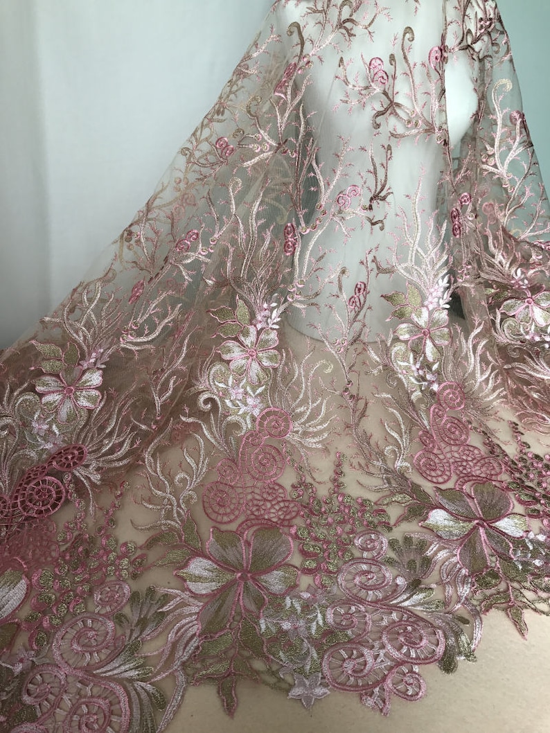 May include: Delicate pink floral embroidered lace fabric. The lace features intricate designs of flowers, leaves, and swirling patterns in shades of pink, white, and gold. The sheer fabric is draped, showcasing the detailed embroidery.