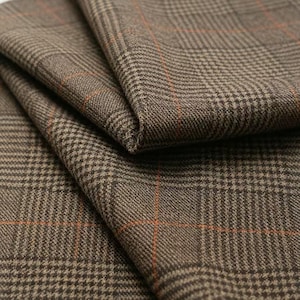 May include: A folded piece of fabric featuring a brown and dark brown glen plaid pattern with thin, intersecting orange lines. The textile has a visible woven texture and is arranged in overlapping layers to show the fabric's drape.