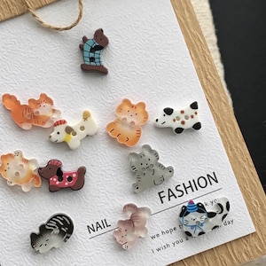 May include: A collection of small, shaped buttons featuring various animal designs, including cats and dogs in colors like orange, gray, brown, and white. Some buttons have patterns like plaid or polka dots. The text NAIL FASHION is printed on the card.