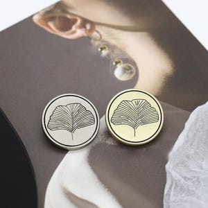 May include: Two round metal pins, one silver and one gold, each featuring a black ginkgo leaf design within a black border. The pins are displayed on a neutral background, showcasing their contrasting colors and intricate details.
