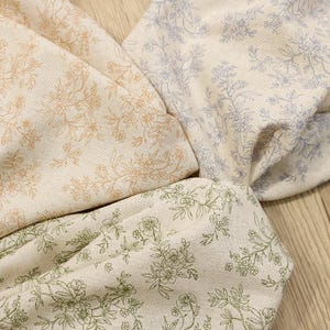 May include: Close-up of four fabric swatches with floral patterns. The fabrics are in shades of beige, with floral designs in peach, blue, and green. The fabric is folded and arranged to show the different patterns and colours.