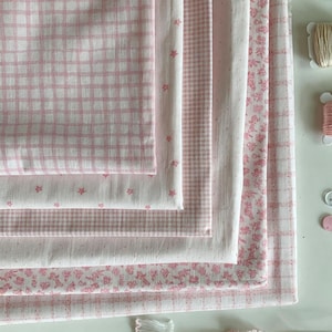 Pode incluir: Uma pilha de quadrados de tecido dobrados com vários padrões em rosa e branco, incluindo xadrez, vichy, pequenas estrelas e estampados florais. Dois pequenos carretéis de linha e dois botões estão posicionados à direita da pilha.