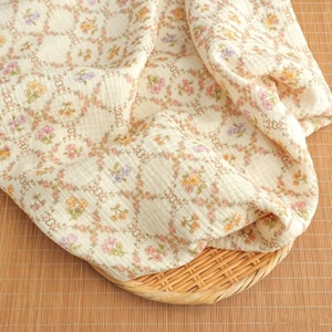 May include: Cream-colored fabric with a floral pattern in shades of pink, yellow, and purple. The fabric is draped over a woven basket. The floral design is arranged in a repeating circular pattern.