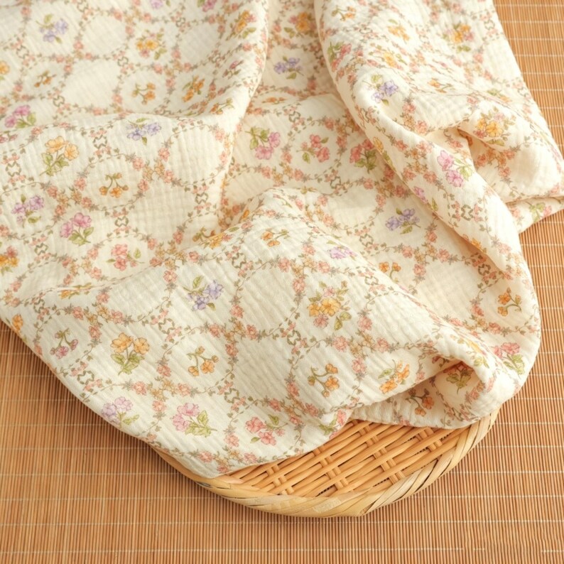 May include: Cream-colored fabric with a floral pattern in shades of pink, yellow, and purple. The fabric is draped over a woven basket. The floral design is arranged in a repeating circular pattern.
