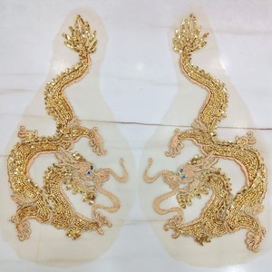 Könnte beinhalten: Zwei goldgestickte Drachenapplikationen. Jeder Drache ist aufwendig mit goldenen Perlen und Pailletten verziert und hat blaue Augen und ein fließendes Design. Die Applikationen befinden sich auf einem durchsichtigen, cremefarbenen Hintergrund.