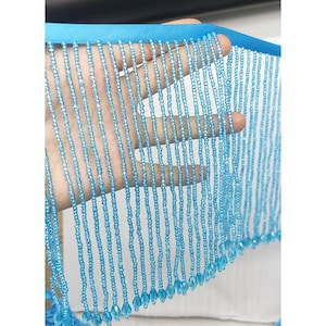 Op de afbeelding: Een lichtblauwe kralenfranje met een effen satijnen lint als bovenrand. De franje bestaat uit talloze verticale strengen van kleine, uniforme lichtblauwe kralen, die elk eindigen in een grotere, druppelvormige kraal aan de onderkant.