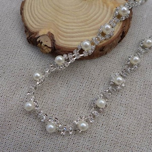 Puede incluir: Una cadena plateada con pequeñas perlas blancas y diamantes de imitación. La cadena está sobre un fondo de tela marrón claro.