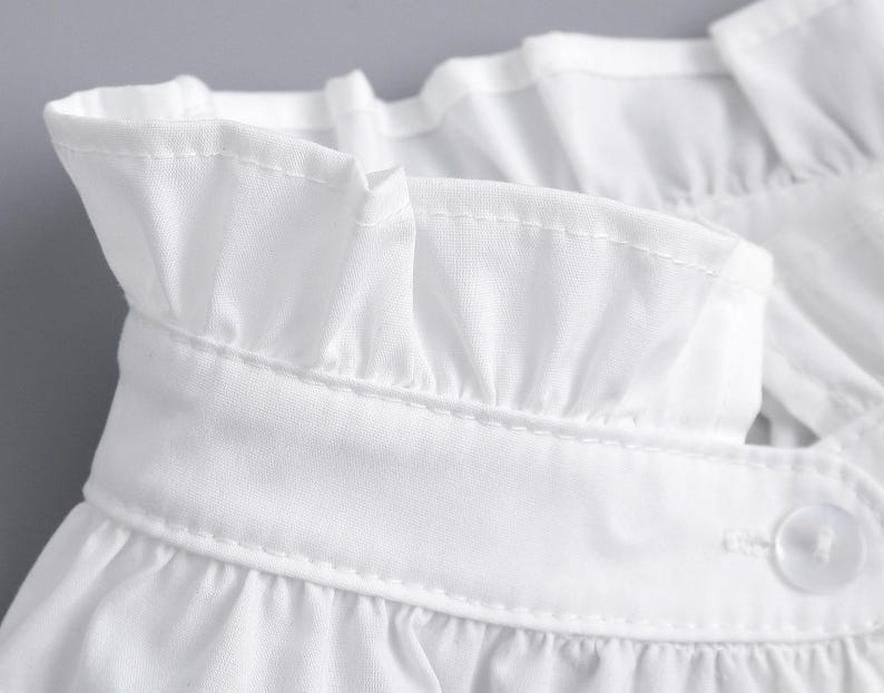 May include: Close-up of a white blouse collar with a ruffled edge. The collar is made of a crisp, lightweight fabric and features delicate stitching. A single white button is visible, adding a touch of detail to the design.