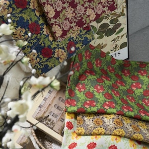 Op de afbeelding: Een assortiment stoffen met bloempatronen in verschillende kleuren, waaronder rood, geel, groen en blauw. De stoffen hebben gedetailleerde bloemontwerpen en zijn aantrekkelijk gerangschikt. Een boek met architecturale afbeeldingen is ook zichtbaar.