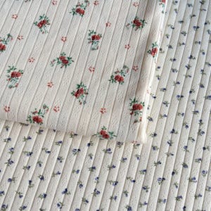 May include: Close-up of two pieces of ribbed fabric. One is cream-colored with a pattern of red roses and green leaves. The other is cream-colored with a pattern of small blue flowers. Both fabrics have a vertical ribbed texture.