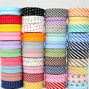 Pode incluir: Quatro pilhas verticais de rolos de fita de tecido com vários padrões, incluindo bolinhas, xadrez vichy, listras diagonais e estrelas. As fitas exibem um amplo espectro de cores como vermelho, azul, amarelo, verde, rosa e preto.