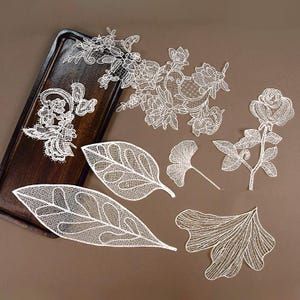 Op de afbeelding: Witte kanten applicaties met bloemen- en bladerenmotieven, gerangschikt op een donkerhouten dienblad. Het fijne kantwerk omvat een roos, diverse bloemen en gedetailleerde bladvormen. De ontwerpen staan tegen een bruine achtergrond.