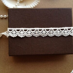 May include: A brown rectangular box wrapped with a white lace trim. The lace has a scalloped edge with floral patterns. A strand of pearls and a seashell are in the background.