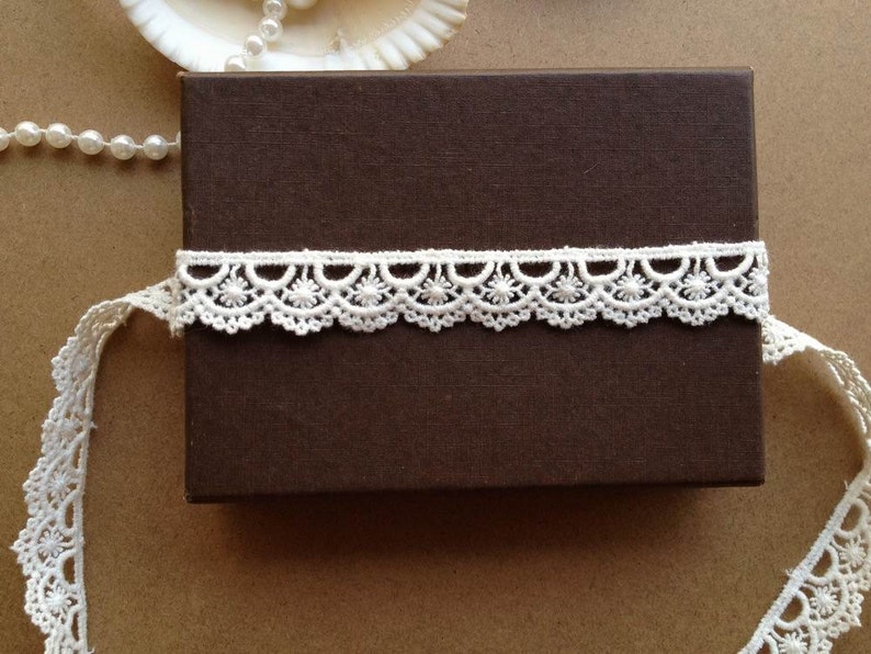 May include: A brown rectangular box wrapped with a white lace trim. The lace has a scalloped edge with floral patterns. A strand of pearls and a seashell are in the background.