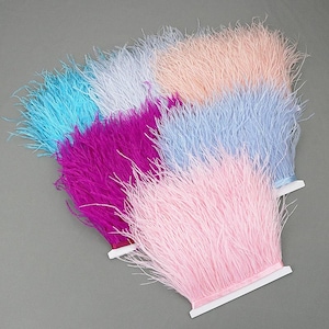 May include: A collection of colorful ostrich feather trim in shades of pink, blue, purple, and peach. Each trim is attached to a white ribbon base, creating a layered effect. The feathers are soft and fluffy.