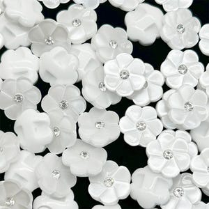 May include: A collection of white flower-shaped embellishments with a central rhinestone. The glossy, petal-like shapes are scattered across a dark background, creating a contrast that highlights the details of each piece. These are likely used for crafts or jewelry making.