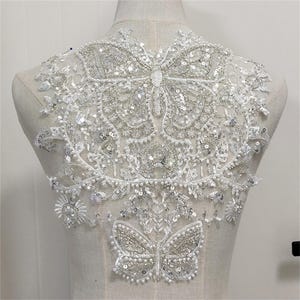 May include: A white lace applique with two silver butterfly designs. The butterflies are embellished with sequins and beads.