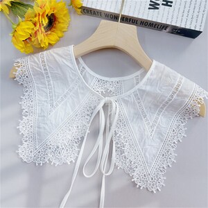 Puede incluir: Un cuello blanco con adornos de encaje y cierre de lazo, exhibido en una percha de madera. El cuello presenta intrincados patrones de encaje floral y geom&eacute;trico. El texto "CREATE WONDERFUL HOMELIFE" es visible en el fondo.