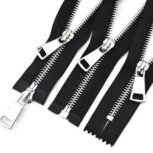 Puede incluir: Primer plano de varias cremalleras negras con tiradores de metal plateado. Las cremalleras están abiertas y cerradas, mostrando los dientes y las lengüetas rectangulares. La imagen está bien iluminada.