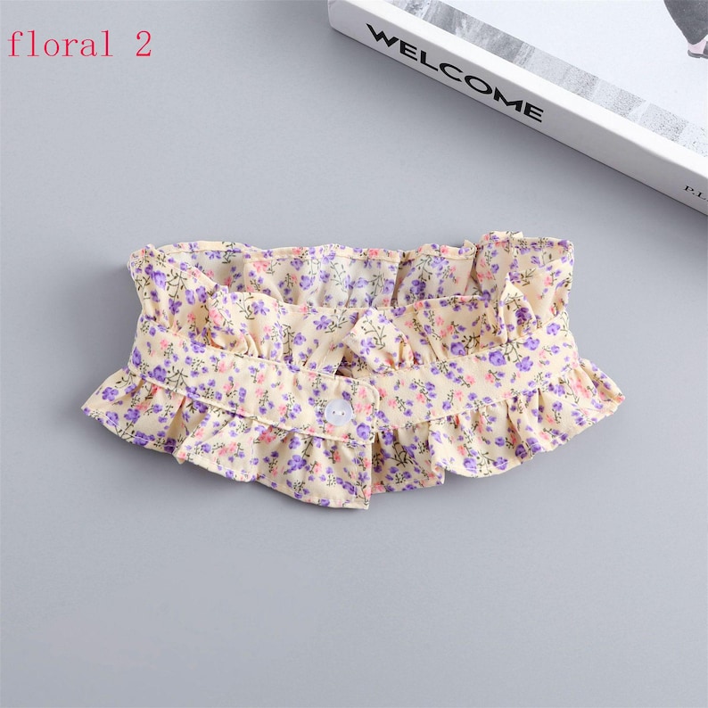 May include: A floral collar with a ruffled edge. The collar is cream-colored with a pattern of small purple and pink flowers. It has a single button closure. The text "floral 2" is in the upper left corner.
