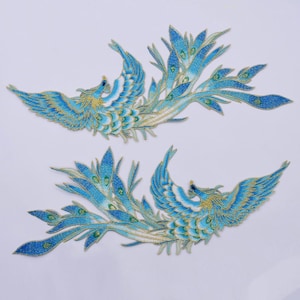 May include: Two embroidered patches featuring a phoenix design. The phoenixes are depicted in shades of blue, with gold and green accents. The patches are detailed with intricate stitching, and are set against a white background.