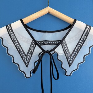 May include: A white sheer collar with black lace trim and a black ribbon tie. The collar has a scalloped edge and is designed to be worn over a shirt or blouse.