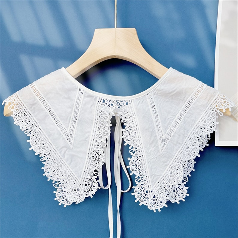 Puede incluir: Un cuello desmontable blanco con ribete de encaje. El cuello presenta un delicado dise&ntilde;o de encaje floral a lo largo de los bordes y un patr&oacute;n en forma de V en el centro. Est&aacute; suspendido de una percha de madera sobre un fondo azul.