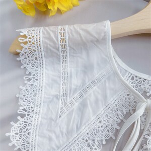 Puede incluir: Cuello de encaje blanco con intrincados patrones florales y geom&eacute;tricos. El cuello presenta una tela delicada y transparente y un cierre de lazo. El dise&ntilde;o incluye un borde festoneado y se muestra en una percha de madera.