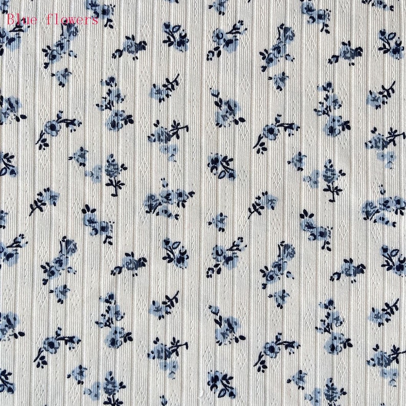 May include: Close-up of a white fabric with a ribbed texture, adorned with a scattered pattern of small blue floral designs. The words "Blue flowers" are visible in the top left corner.