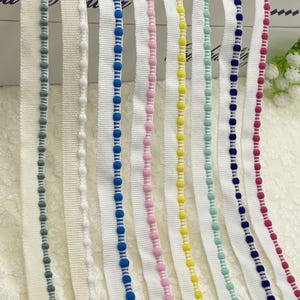May include: Close-up of several ribbons with a white base and colorful beaded trim. Colors include teal, white, blue, pink, yellow, and red. The ribbons are arranged vertically, showcasing the variety of colors and textures.