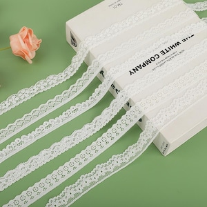 May include: Close-up of several rolls of white lace ribbon with intricate floral and geometric designs. The lace is laid out on a white book with the text "THE WHITE COMPANY". A peach rose is in the background. The lace is perfect for crafts.