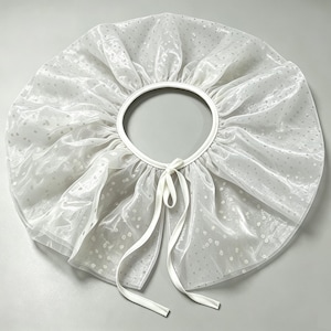 May include: A white sheer ruffled collar featuring a subtle polka dot pattern throughout the fabric. The circular accessory includes a solid white trim along the inner neckline and two long white ribbon ties for fastening at the front.