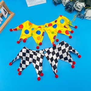 Puede incluir: Dos cuellos de bufón hechos de tela satinada. Un cuello es de color amarillo brillante con lunares multicolores, y el otro es de cuadros blancos y negros. Ambos cuellos tienen bordes puntiagudos, cada uno con un pequeño pompón rojo redondo.