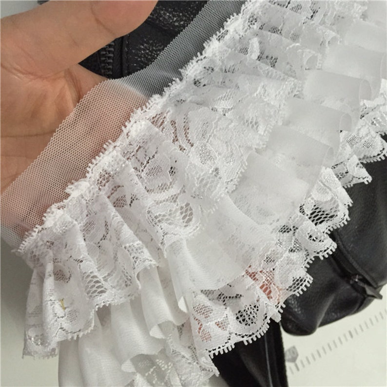 May include: White lace trim with a ruffled edge and a sheer mesh backing. The lace is delicate and intricate, with a floral pattern.