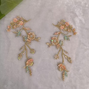 Puede incluir: Dos apliques florales a juego con lentejuelas color melocotón y rosa claro, cuentas iridiscentes y bordados en hilo metálico dorado. El diseño consiste en tallos ramificados con pequeñas hojas y flores dispuestas en un patrón de enredadera.