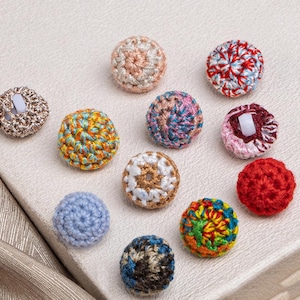 Puede incluir: Una colección de botones de ganchillo hechos a mano en varios colores y patrones. Los botones son redondos y presentan intrincados trabajos de puntada, con colores como azul, rojo, amarillo y rosa. Algunos botones tienen un vástago en la parte posterior para coser.
