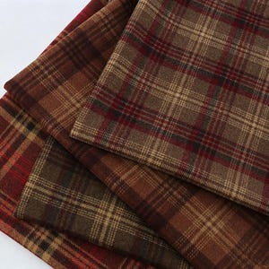 Puede incluir: Una colección de piezas de tela dobladas con un patrón de cuadros clásico en tonos marrón, beige y rojo oscuro. El tejido muestra una textura tejida con líneas horizontales y verticales que se cruzan formando un diseño de cuadrícula constante.
