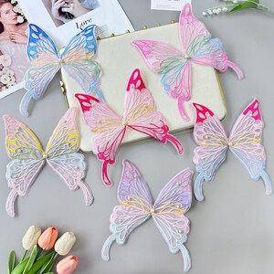 May include: A selection of butterfly-shaped appliques in various colours. The sheer wings are in shades of pink, blue, yellow, and purple, with white stitching and small pearl details. The appliques are displayed on a light grey surface.