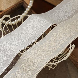 May include: A wide strip of off-white sheer fabric featuring intricate white floral embroidery. The design consists of repeating circular flower motifs surrounded by delicate leaf patterns, with a finished edge along both sides of the ribbon.