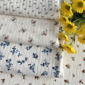 May include: Close-up of three rolls of white ribbed fabric with floral patterns. The fabric features small, delicate flowers in shades of blue, red, and pink. The fabric is soft and has a textured surface. Yellow flowers are in the background.