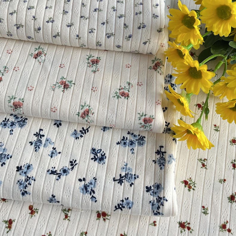 May include: Close-up of three rolls of white ribbed fabric with floral patterns. The fabric features small, delicate flowers in shades of blue, red, and pink. The fabric is soft and has a textured surface. Yellow flowers are in the background.