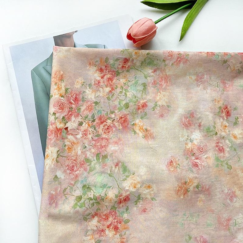 May include: A floral fabric featuring a pattern of pink, peach, and yellow roses with green leaves. The roses are clustered together, creating a romantic and vintage aesthetic. The fabric is draped over a surface, with a pink tulip in the upper right corner.