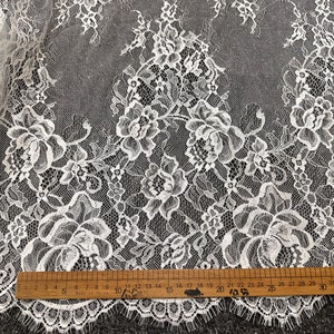 May include: White lace fabric with a floral pattern. The lace has a scalloped edge and is measured with a ruler in inches.