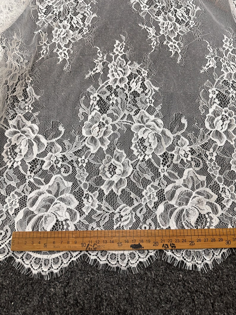 May include: White lace fabric with a floral pattern. The lace has a scalloped edge and is measured with a ruler in inches.