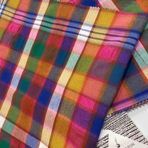Puede incluir: Una tela tejida que presenta un vibrante patrón de cuadros multicolor con líneas que se cruzan en tonos rosa, naranja, amarillo, verde, azul y morado. El textil muestra un diseño de cuadrícula con anchos de rayas y bloques de color.