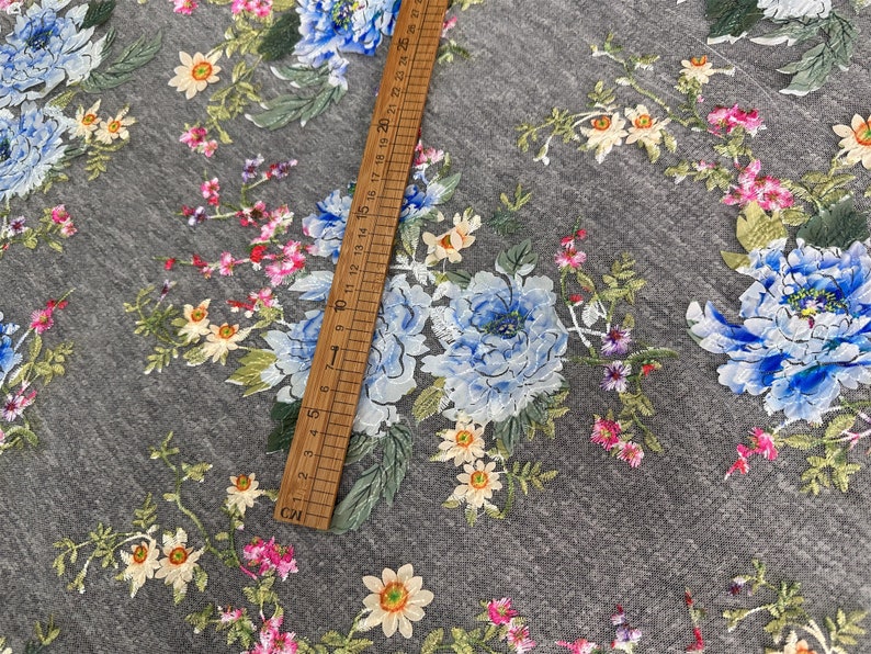 Puede incluir: Tela de tul gris con un patr&oacute;n floral con flores azules, rosas y amarillas. La tela se mide con una regla en pulgadas.