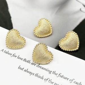 May include: Four gold-colored heart-shaped embellishments. Each heart has a textured, ridged surface and a beaded border. The hearts are arranged on a white surface with text that reads "A taboo for love: Both are dreaming the future of each but always think of the..."