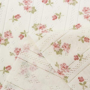 May include: A cream-colored fabric featuring a repeating pattern of small pink roses with green leaves. The textile has a delicate, open-knit texture with vertical rows of tiny eyelet perforations running throughout the material's surface.
